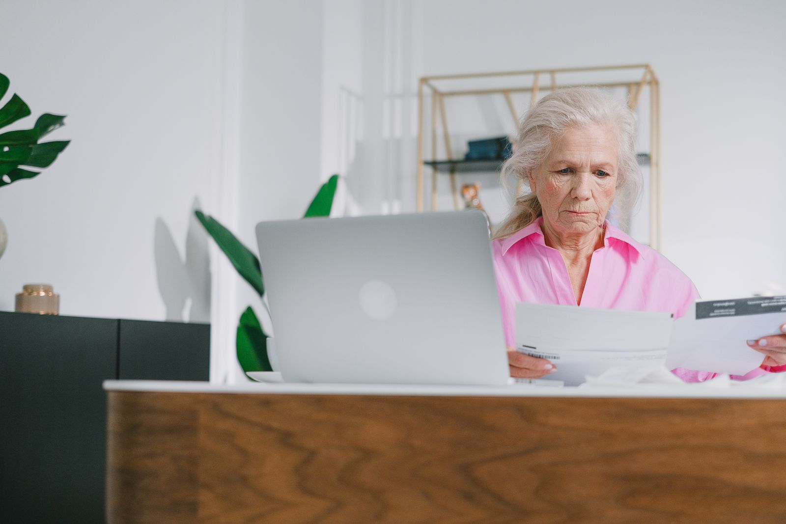 A grey-haired woman reading papers at her desk beside a laptop